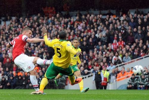 Arsenal's Lukas Podolski (L) scores a goal as Norwich City's Sebastien Bassong (C) ties to block, April 13, 2013